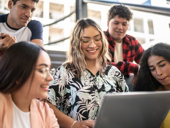 1455041863 Gruppe von Studierenden sitzt gemeinsam auf Stufen und blicken auf den Laptop einer Kollegin
